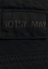 Tessuto di denim nero con cuciture visibili; etichetta rettangolare non decorata che mostra il testo "NOISY MAY" in un carattere semplice.