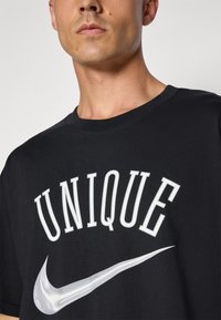 Közeli kép egy személyről, aki fekete pólót visel, rajta fehér nagybetűs "UNIQUE" felirattal, alatta pedig egy torzított fehér Nike logóval.