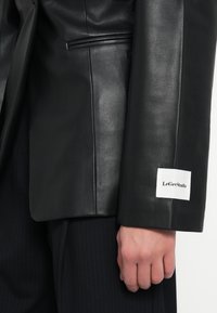 Čierny kožený blejzer s klopovým golierom, bočným vreckom a bielou štítkom s nápisom "LeGerStudio" na manžete. Textúrovaný povrch.