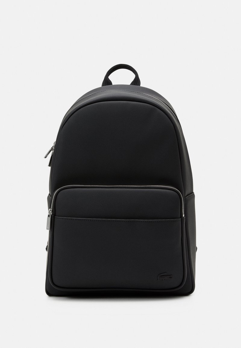 Sac à dos noir texturé avec poche avant zippée, poignée supérieure et petit logo embossé dans le coin inférieur droit sur un fond clair.