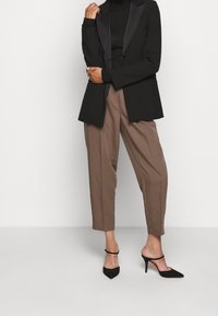 Frau in braunen, schmal geschnittenen Hosen, schwarzen High Heels, schwarzem Rollkragenpullover und schwarzem Blazer vor einem schlichten weißen Hintergrund.