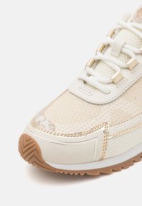 Zapatilla beige claro con parte superior de malla, detalles en dorado y cordones blancos. Suela texturizada con agarre de goma marrón. Presenta costuras decorativas.
