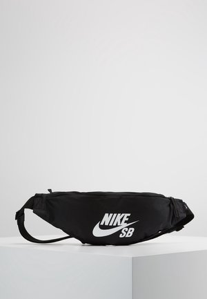 Riñonera negra Nike SB con logo blanco, correa ajustable y compartimentos con cremallera, exhibida sobre un pedestal blanco.