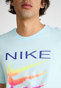 Šviesiai mėlyna Nike marškinėliai su ryškiu tamsiai mėlynu logotipu ir spalvotu gradientu swoosh dizainu, kuriame dominuoja rožinė, geltona ir žalia. Trumpi rankoviai.