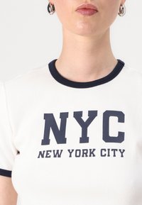 Bijela rebrasta majica s mornarskom plavom obrubom, s odvažnim tekstom "NYC" i "NEW YORK CITY" u tamnoplavoj boji. Kratak rukav i prilagođen dizajn.