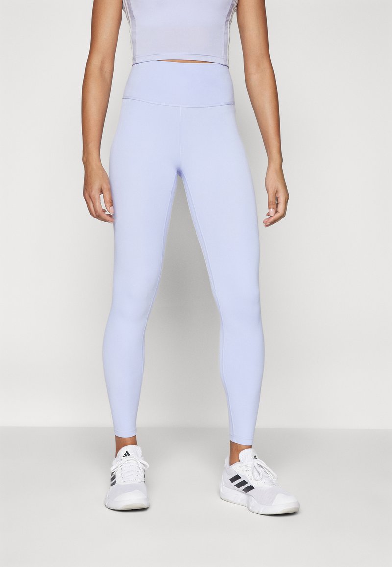 Világoslila leggings, magas derékkal és sima, rugalmas textúrával. Fehér sportcipőkkel párosítva, fekete csíkokkal.