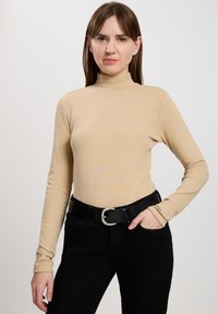 Beżowy sweter z długim rękawem i golfem uszyty z miękkiego materiału, w połączeniu z czarnymi dżinsami z wysokim stanem i czarnym paskiem z srebrną klamerką.