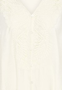 Blouse blanche avec motifs floraux en dentelle autour du col en V et fermeture à boutons sur le devant, avec des détails de broderie délicats et complexes.