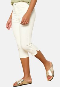 Pantalones recortados blancos con un diseño ajustado, que cuentan con acentos de corte en el dobladillo. Combinados con sandalias deslizantes metálicas en dorado sobre una suela texturizada.