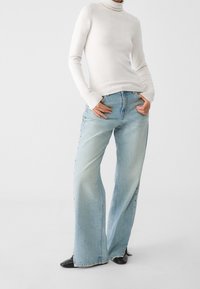 Jeans de tiro alto de color azul claro con textura deslavada, con botones en los laterales. Combinados con un suéter ceñido blanco de cuello alto. Zapatos negros de punta. 