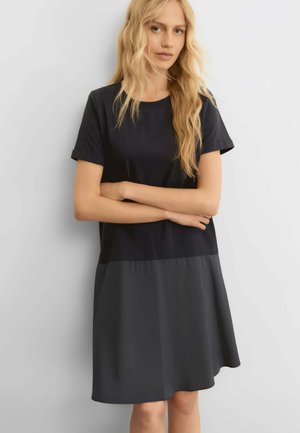 Giovane donna con lunghi capelli biondi in piedi con le braccia incrociate, che indossa un vestito a blocchi di colore nero e grigio scuro a maniche corte.