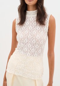 Kanten blouse met hoge hals, met een witte bovenkant en een lichtere crèmekleurige onderkant, die een bloemenpatroon en een scalloped zoom toont.