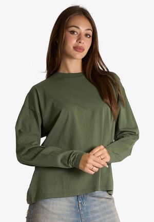 Donna con lunghi capelli castani, indossa una camicia verde oliva a maniche lunghe larga e jeans azzurri chiari, in piedi con le mani giunte davanti.
