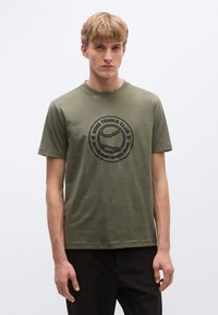 Olívazöld pamut póló kerek nyakkivágással és rövid ujjakkal. Középen egy fekete teniszlabda grafika "BOSS TENNIS CLUB" felirattal.