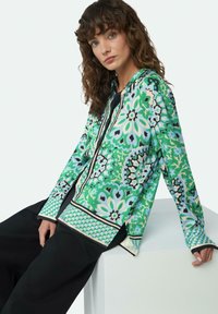 Langärmliges Hemd mit Knöpfen, das ein lebhaftes grünes Blumenmuster aufweist, mit schwarzem Besatz und einer glatten Stofftextur, kombiniert mit schwarzen Hosen.