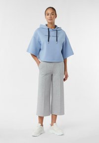 Cappuccio corto azzurro chiaro con maniche ampie e cordini neri, abbinato a pantaloni grigi a gamba larga. Il modello indossa sneakers bianche.