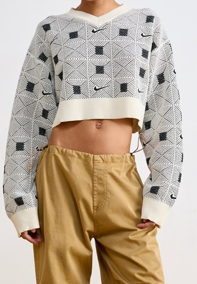 Pull tricoté blanc et noir à motif, court avec manches longues, associé à un pantalon beige ample, laissant voir le ventre et le nombril.