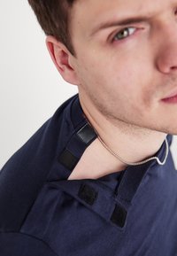 Tengerészkék póló, lekerekített nyakkivágással, a vállaknál tépőzáras pántokkal, amelyek lehetővé teszik a méret állítását, valamint egy ezüstlánc látható a nyaknál.