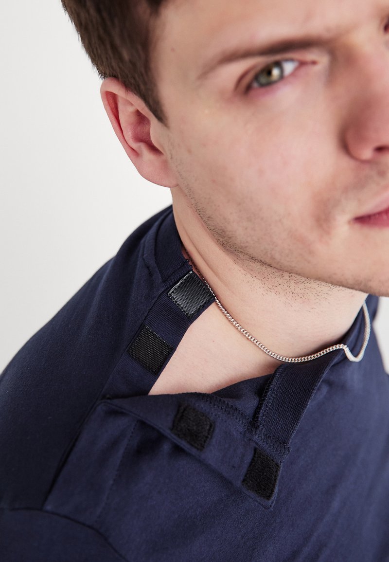 Tengerészkék póló, lekerekített nyakkivágással, a vállaknál tépőzáras pántokkal, amelyek lehetővé teszik a méret állítását, valamint egy ezüstlánc látható a nyaknál.