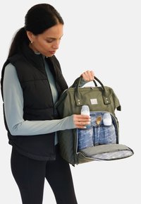 Bolsa de pañales verde con textura acolchada, que presenta compartimentos interiores grises que sostienen biberones y una cuchara. Doble asa superior para llevarla.