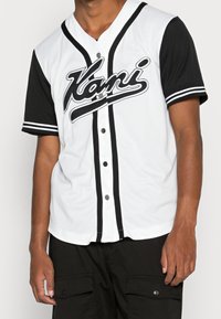 Bílý baseballový dres s černými krátkými rukávy, na kterém je velké logo "Kani", černé detaily a zapínání na knoflíky vpředu.