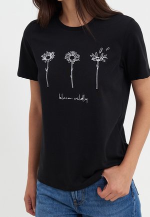 Sort t-shirt med tre hvide tusindfryd og teksten "bloom wildly," båret med blå jeans, hånd i lommen.