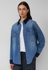 Camicia in denim blu medio con colletto in corduroy scuro, bottoni a pressione e due tasche sul petto; vestibilità rilassata, maniche lunghe con polsini a coste.