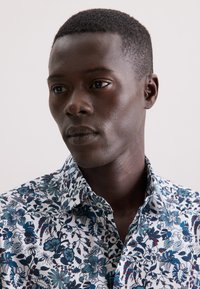 Camicia bianca con stampa blu e floreale, caratterizzata da un colletto a punta e bottoni. Il tessuto sembra liscio con intricati motivi floreali.