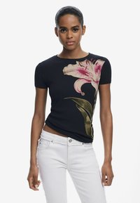Zwarte geribde T-shirt met een grote roze lelieprint en groene bladeren aan één kant. Gecombineerd met witte jeans die een getailleerd ontwerp tonen.