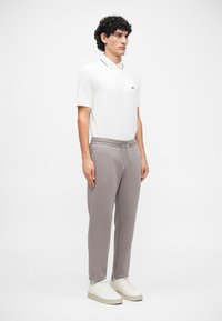Cămașă polo albă cu guler negru și logo, purtată cu pantaloni de trening gri deschis, cu talie elastică, și sneakers albi.