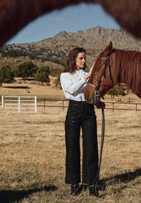 Zwarte, wijde broek met borduursels, gecombineerd met een wit shirt met lange mouwen. Een bruine paard wordt geaaid in een grasrijke buitenomgeving.