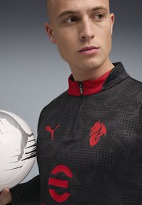 Haut de sport zippé noir avec col rouge et accents, présentant un motif texturé, un logo, et tenant un ballon de football blanc avec des détails gris.