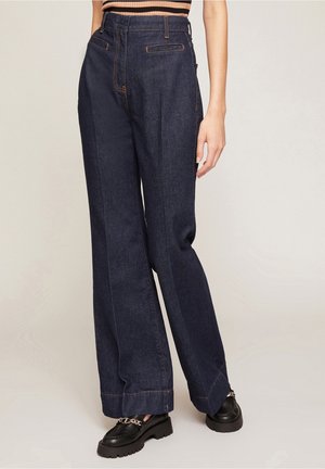 Femme portant un jean bleu foncé taille haute à jambes larges avec des coutures visibles à l'avant et des mocassins noirs épais avec un détail chaîne.