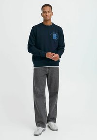 Marinefarbener Sweatshirt mit einem blauen "B"-Logo, Rundhalsausschnitt, langen Ärmeln, kombiniert mit grauen weiten Jeans und weißen Sneakers.