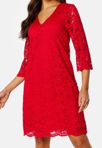 Robe en dentelle rouge avec un décolleté en V, des manches trois-quarts, des motifs floraux et un ourlet festonné, fabriquée à partir d'un tissu léger et texturé.