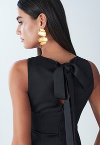 Black satin dress features a back tie detail and a hidden zipper. Gold statement earrings in a textured leaf design.