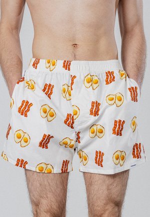 Uomo che indossa boxer bianchi con stampa di uova fritte e strisce di pancetta, in piedi con le mani nelle tasche su sfondo semplice.