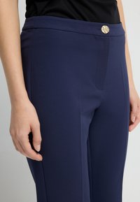 Marinblå byxor med en slät textur, figurnära design och en synlig guldknytned vid midjan, som visar en ren och enkel estetik.