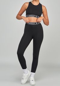 En person bär en svart atletisk crop top och leggings med Siksilk-logotypen, poserar självklart i vita sneakers och strumpor mot en grå bakgrund.