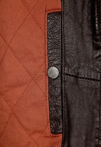 Tikkikuvioitu ruosteenvärinen kangas ja musta nahkainen paneeli hopeanvärisellä nepparilla. Nahalla on teksturoitu pinta ja näkyvät ompelusaumat.