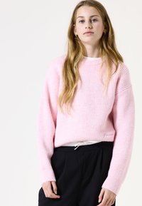 Rosa gerippter Strickpullover mit langen Ärmeln, runden Halsausschnitt und lockerer Passform. Getragen über schwarzen Hosen mit weißen Kordelzugdetails.