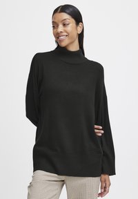 Pulover negru cu guler înalt, cu o croială lejeră și manșete striate, asortat cu o pereche de pantaloni bej cu carouri. Textură moale și design casual.
