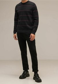 Schwarzer Pullover mit kastanienbraunen Streifen, Rundhalsausschnitt und gerippten Bündchen, kombiniert mit schwarzen, taillierten Hosen und klobigen schwarzen Schuhen.