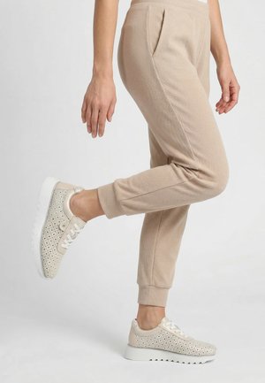 Person iført beige ribbet joggingbukser og cremefarvede perforerede sneakers, løfter det ene ben bagud mod en ensfarvet hvid baggrund.