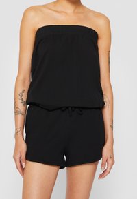 Donna che indossa un romper senza spalline nero con una parte superiore elasticizzata e vita con cordoncino, mostrando tatuaggi su entrambe le braccia su uno sfondo semplice.