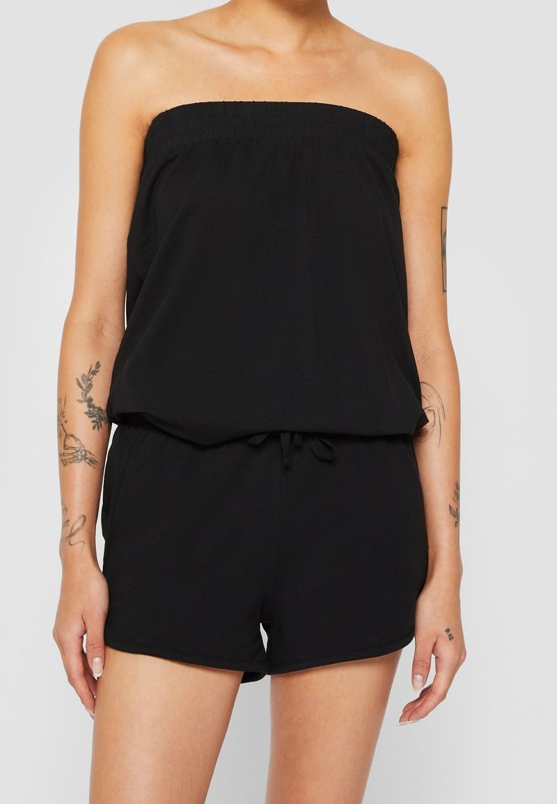 Donna che indossa un romper senza spalline nero con una parte superiore elasticizzata e vita con cordoncino, mostrando tatuaggi su entrambe le braccia su uno sfondo semplice.