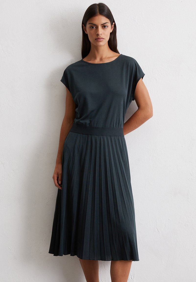Abito plissettato in verde scuro con una vestibilità rilassata, maniche a cappuccio e vita a costine. Realizzato in un tessuto morbido e liscio con una silhouette fluida.