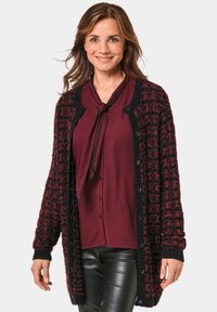 Langer Cardigan aus burgunderfarbenem und schwarzem strukturiertem Strick, mit Knopfverschluss und gerippten Bündchen, über einem burgunderfarbenen Top mit Schleifendetail getragen.