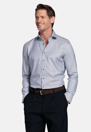 Lachende man met handen in de zakken draagt een klein geruit blauw-wit overhemd, donkere broek en een bruine riem tegen een effen achtergrond.