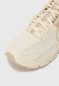 Tenisice bež barve z mrežastim zgornjim delom, teksturiranimi poudarki in bež prekrivali. Šnire so svetlo rjave barve s subtilnimi sivo-rjavimi črtami.